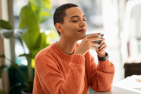 a young woman in a cozy orange sweater closes eyes smelling her mug of coffee