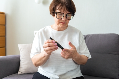 older woman wearing glasses checks her blood glucose at home on her couch