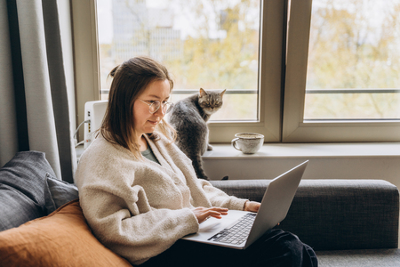 Woman on the couch with her laptop while her cat sits in a windowsill
