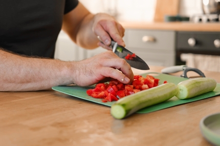 A close up of a man's hands as he dices bell peppers and cucumbers on a cutting board in the kitchen