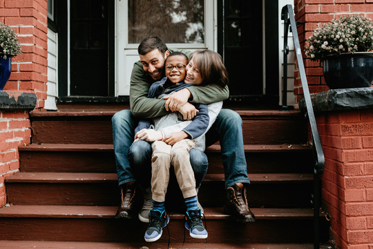 Mom and dad laughing and hugging child sitting on front porch steps of home