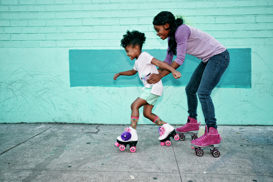 mother teaching daughter to roller skate in front of blue brick wall