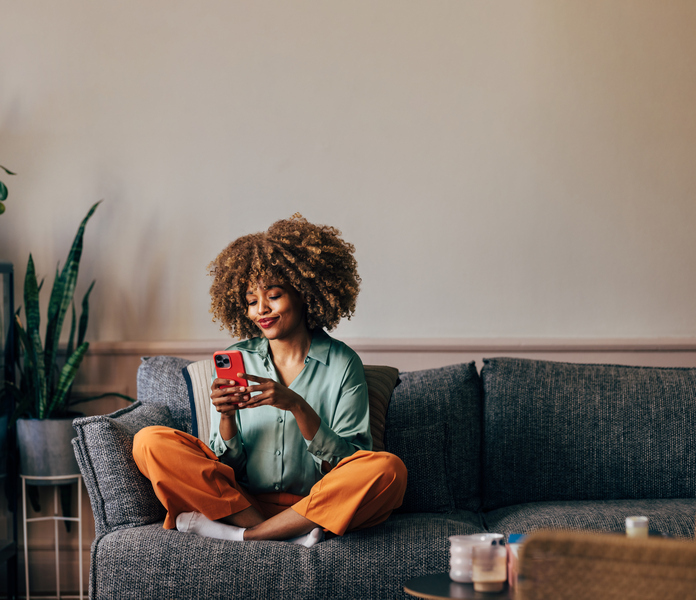 A young woman with curly hair sitting cross-legged on a couch, looking at her phone