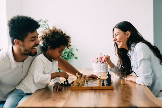 Parents laughing while teaching daughter to play chess at kitchen table