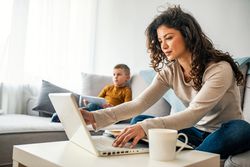 mom working on laptop with son on couch in the background