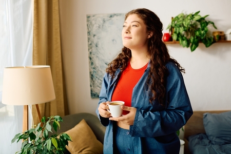 a middle-aged woman in a denim jacket sips coffee as she gazes out her living room window