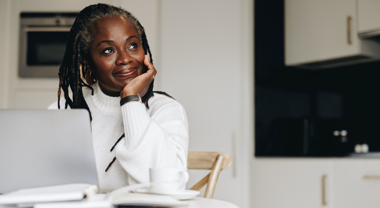 a woman sitting at the kitchen table on the computer looking off into the distance with a smile.