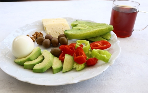 A plate of cheese, olives, nuts, avocados, cucumbers, peppers and an egg on a plate next to a cup of tea