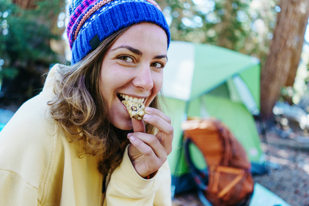 college-age girl in a yellow hoodie and blue beanie eating a granola bar in front of green tent at campsite