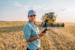 woman with work tablet standing in farmland with large tractor behind her