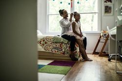 Sitting on edge of a bed, mom arranging hair of daughter who is dressed as a ballerina. 