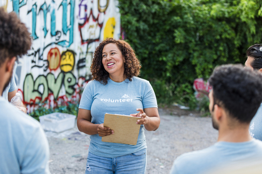 woman in volunteer tee holding clipboard directing group near graffittied wall
