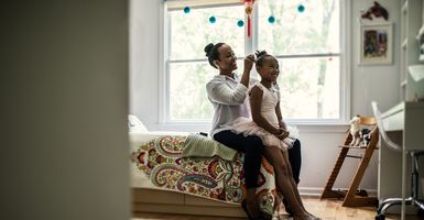 mom sitting on bed doing ballerina daughter's hair