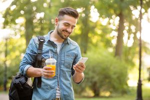 Man walking through park with backpack and juice looking at smartphone