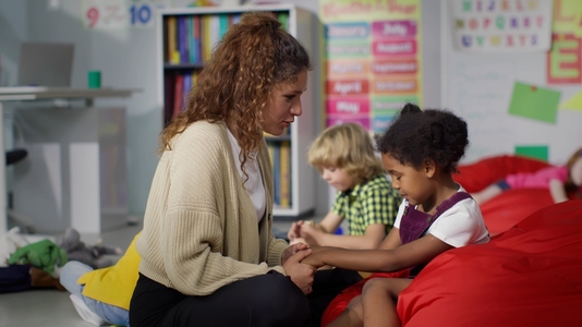 younger female preschool teacher holding female student's hand consoling her