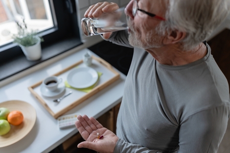 and older man taking medications drinks a glass of water in his kitchen