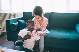 young couple in living room using smartphone together while woman reads