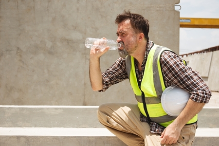 middle-aged male utility worker in yellow vest drinks water in summer heat