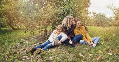 mom and two daughters sitting in orchard under apple tree