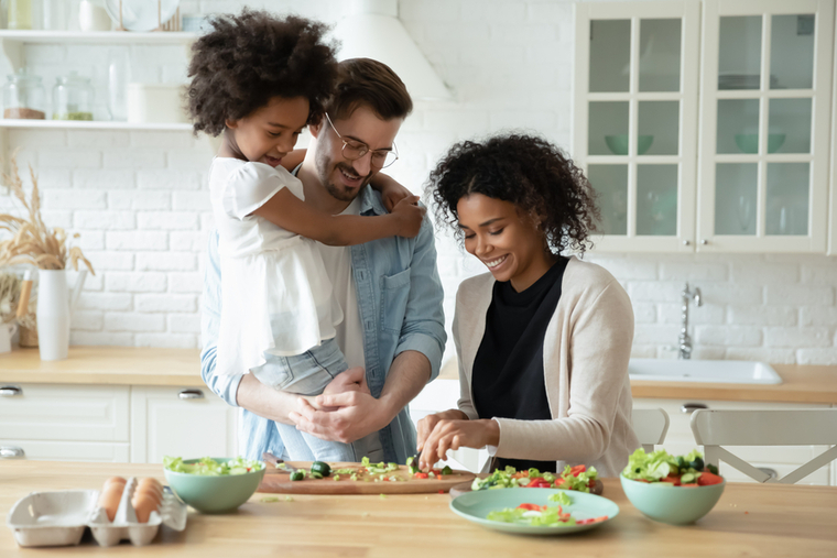 Dad holding daughter in kitchen while mom chops vegetables for salad