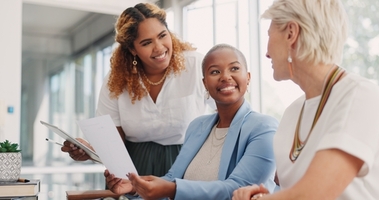 three female coworkers seated at a desk reviewing documents in a sunny office. 