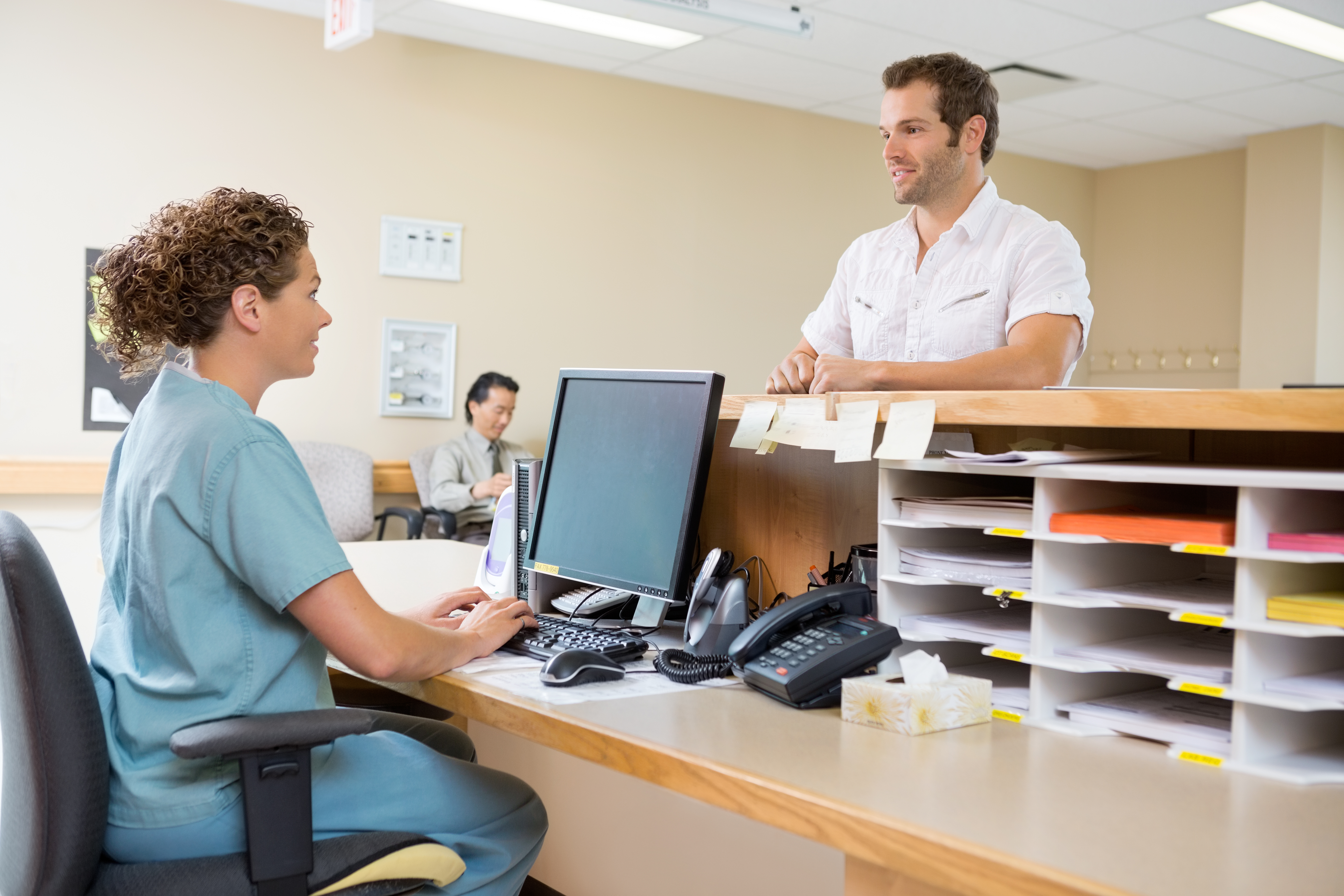 healthcare worker helping patient in doctor office at desk with computer and phones.