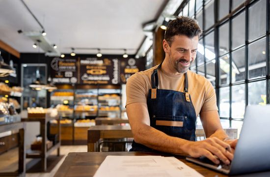 man in apron working on laptop and paper in bakery