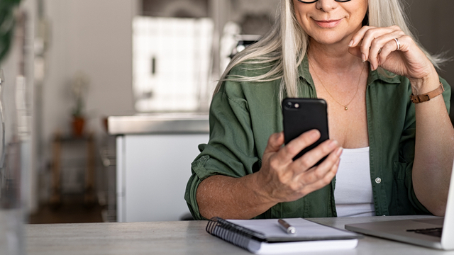 gray-haired woman smiling at phone in front of laptop sitting in kitchen